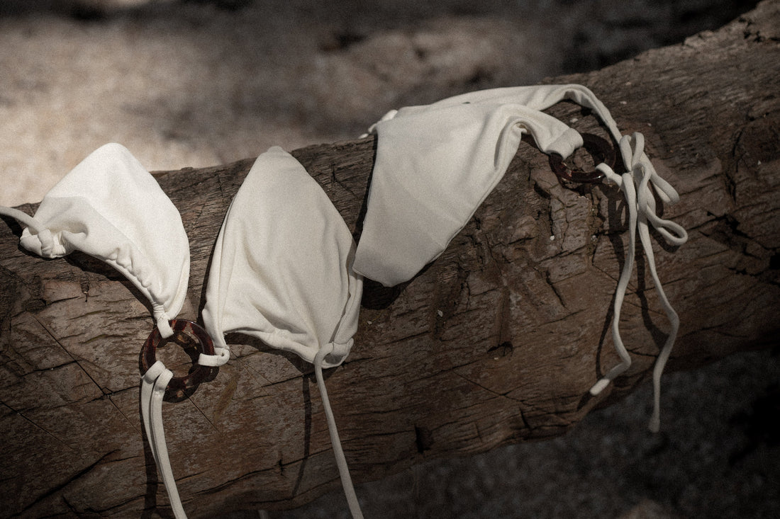 Trois hauts de maillot de bain blancs noués, posés sur une branche en bois dans un décor naturel